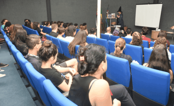 Participantes estão sentados em auditório voltado para o palco onde uma pessoa está vestida de bruxo