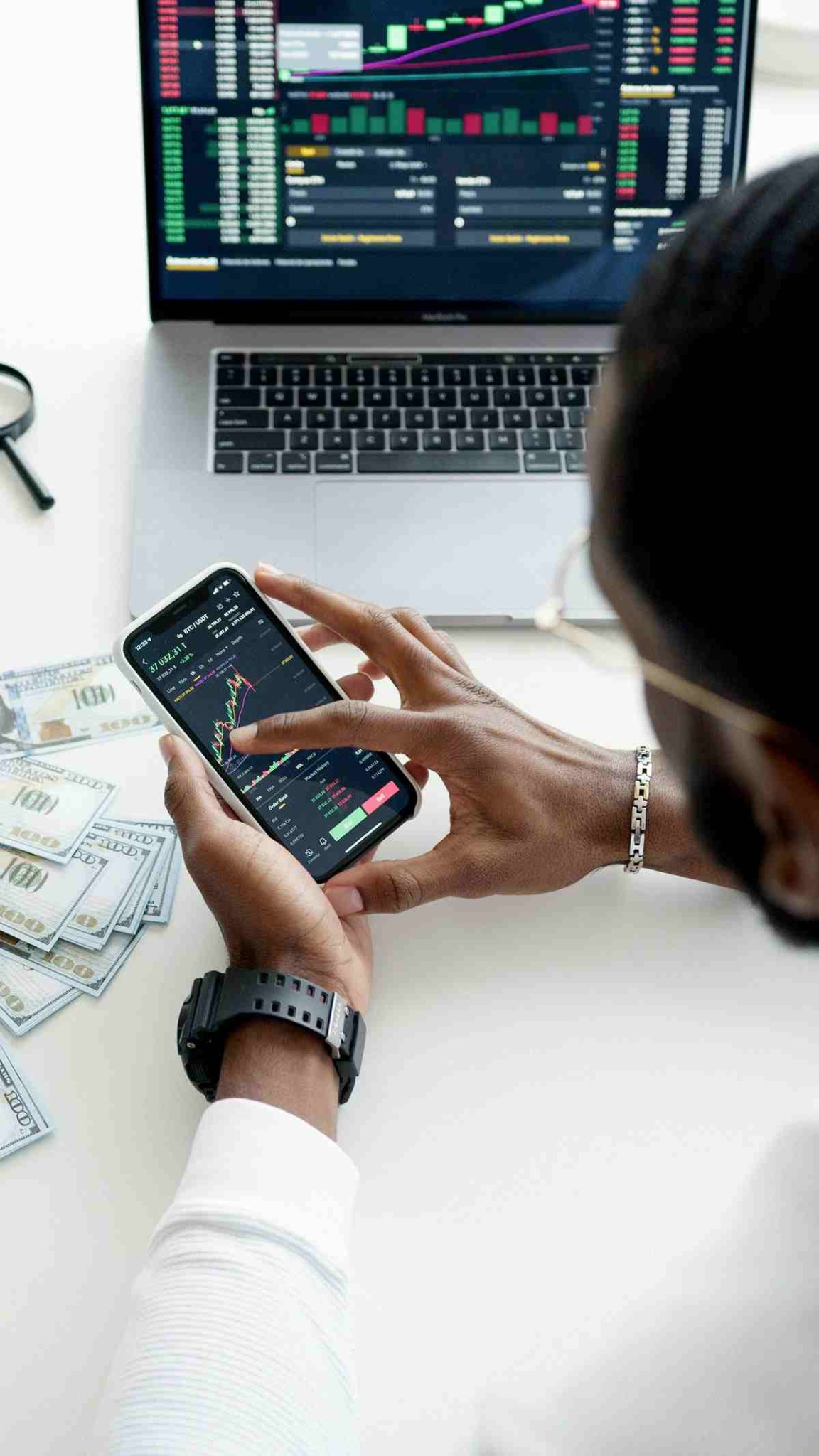 Uma pessoa analisando gráficos de ações ou criptomoedas Bitcoin em um smartphone e laptop, com notas de dólar na mesa. Uma pessoa analisando gráficos de ações ou criptomoedas Bitcoin em um smartphone e laptop, com notas de dólar na mesa.