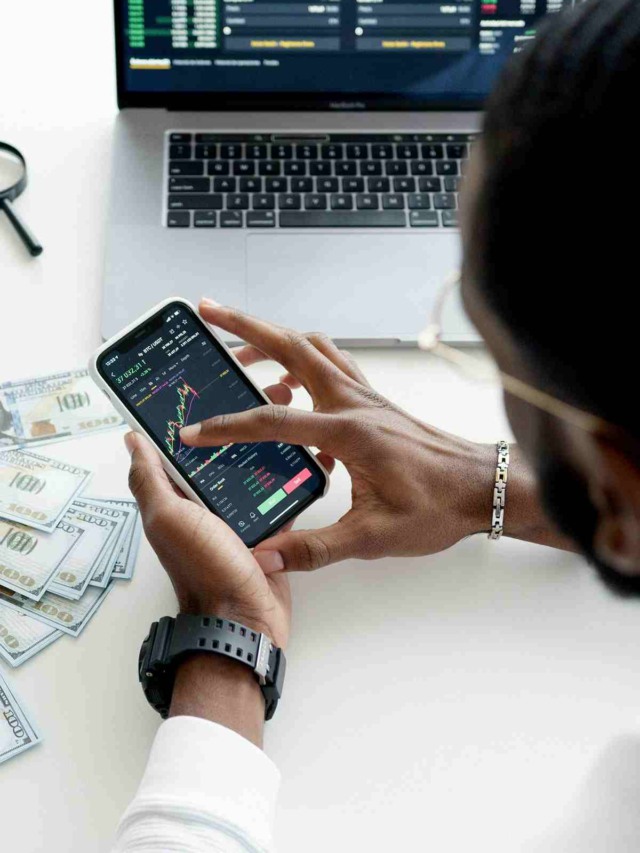 Uma pessoa analisando gráficos de ações ou criptomoedas Bitcoin em um smartphone e laptop, com notas de dólar na mesa.