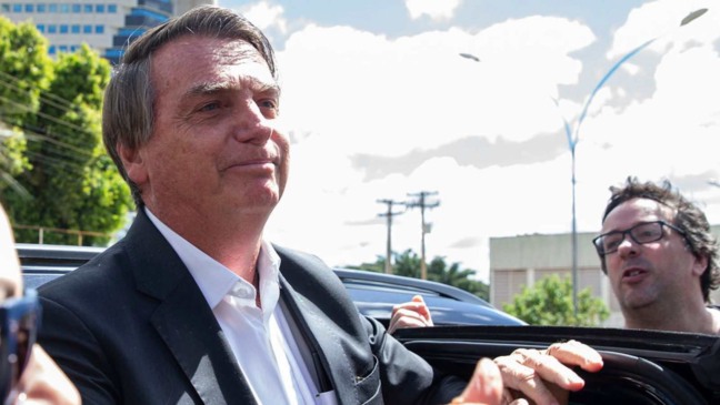 Ex-presidente Jair Bolsonaro de terno preto e camisa branca, ao lado de um veículo, em ambiente externo com céu nublado ao fundo.