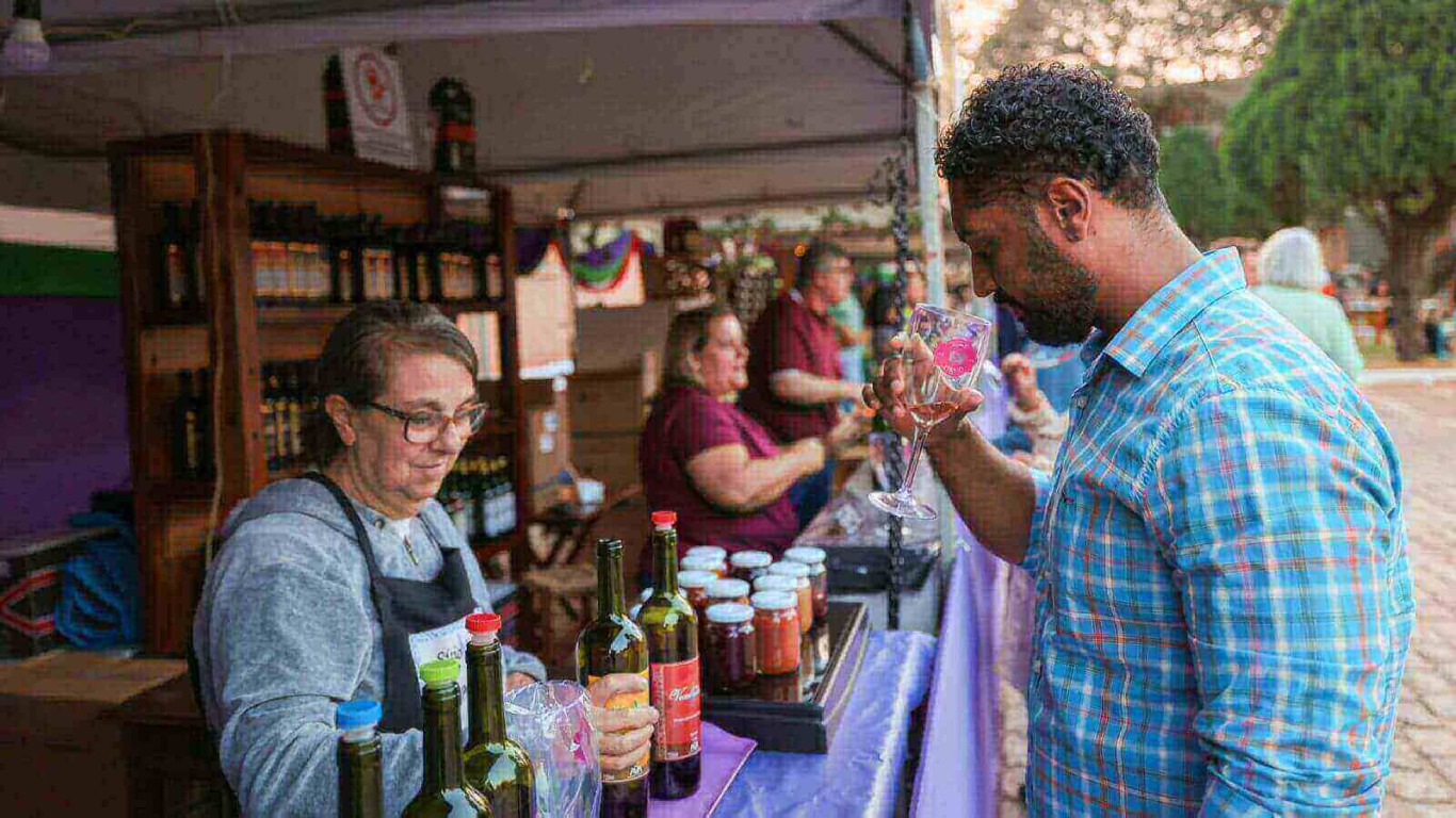 Homem degustando vinho em um estande de feira de vinhos ao ar livre, com várias garrafas e produtos expostos.