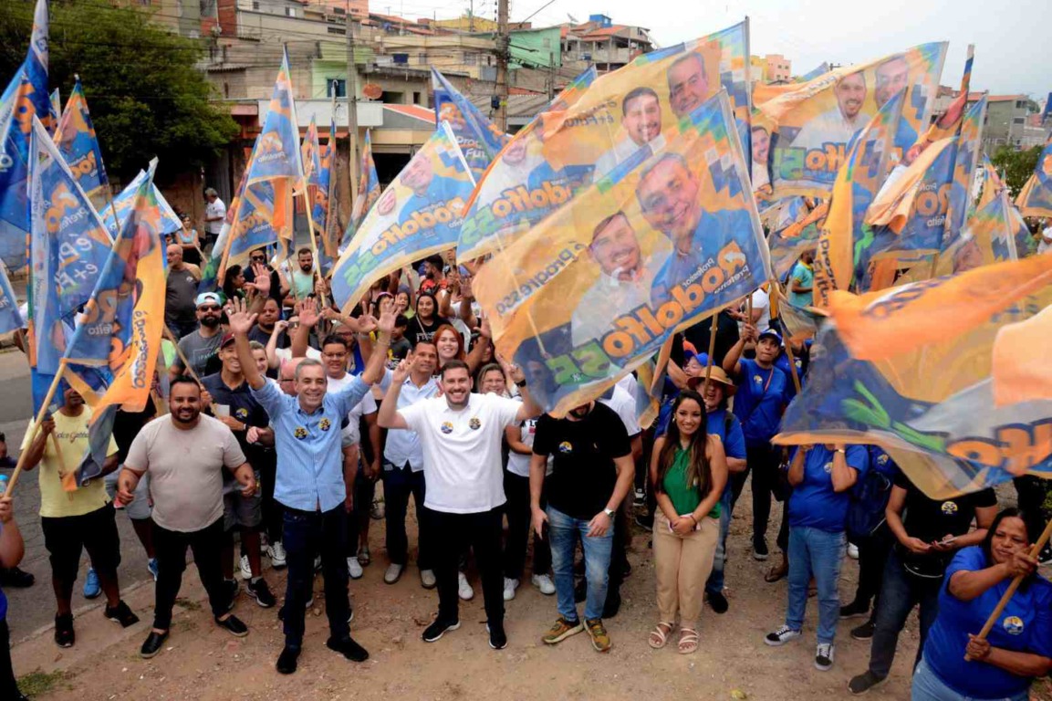 Professor Rodolfo, cercado por apoiadores, lidera caminhada eleitoral em Várzea Paulista, com bandeiras de campanha ao fundo.