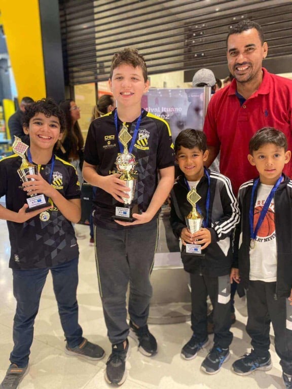 Grupo de crianças e um adulto sorrindo com troféus e medalhas, comemorando uma vitória em um ambiente de competição.
