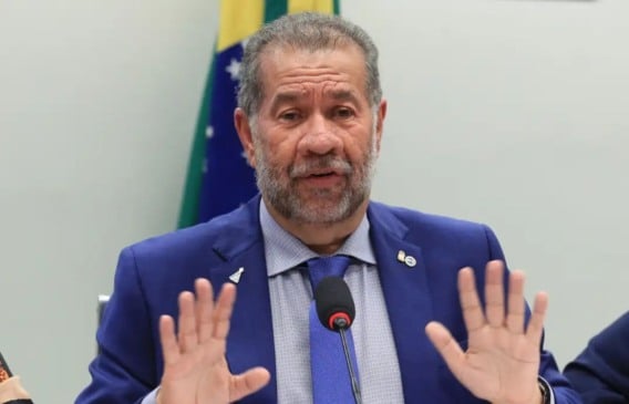 Carlos Lupi, de terno azul e gravata, gesticula com as mãos em microfone, com bandeira do Brasil desfocada ao fundo.