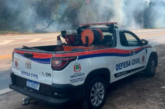Imagem de uma caminhonete da Defesa Civil de Várzea Paulista estacionada ao lado de uma rodovia em Jundiaí, com fumaça de incêndio ao fundo.