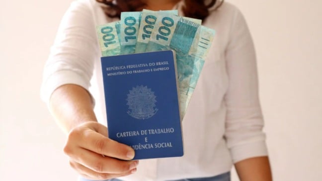 Mulher segurando carteira de trabalho e cédulas de 100 reais.