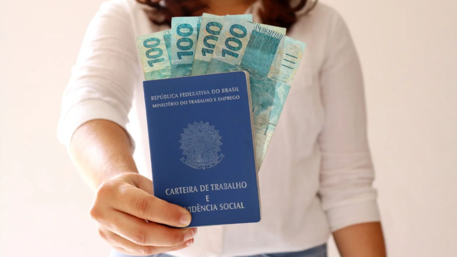Mulher segurando carteira de trabalho e cédulas de 100 reais.