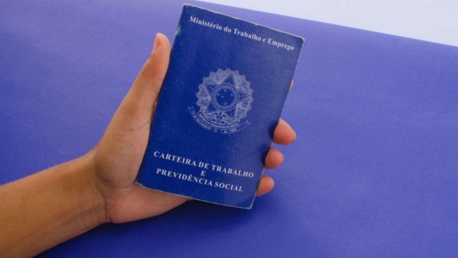 Mão segurando Carteira de Trabalho e Previdência Social azul sobre fundo roxo.