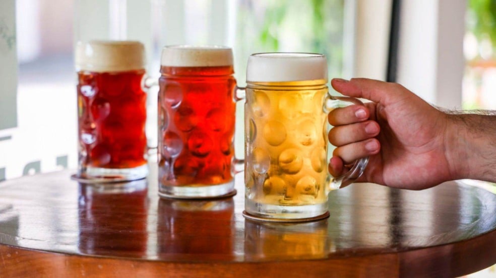 Canecas de chope artesanal, em tons âmbar e dourado, sobre mesa de madeira; uma mão segura uma das canecas.
