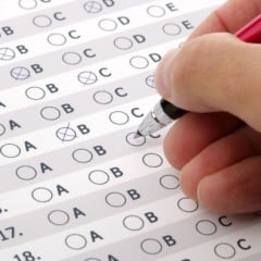 Mão preenchendo cartão-resposta de exame de múltipla escolha com caneta.