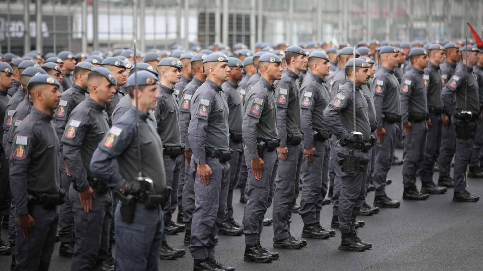 Grupo de policiais militares fardados em formação, perfilados lado a lado, durante solenidade ou treinamento em via pública.