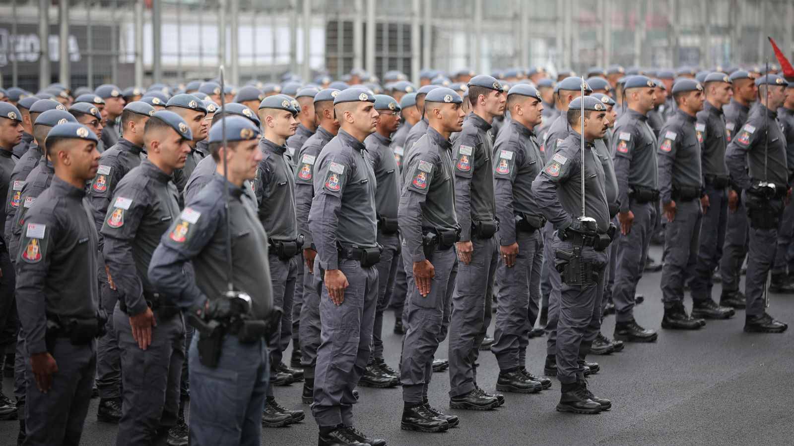 Grupo de policiais militares fardados em formação, perfilados lado a lado, durante solenidade ou treinamento em via pública.