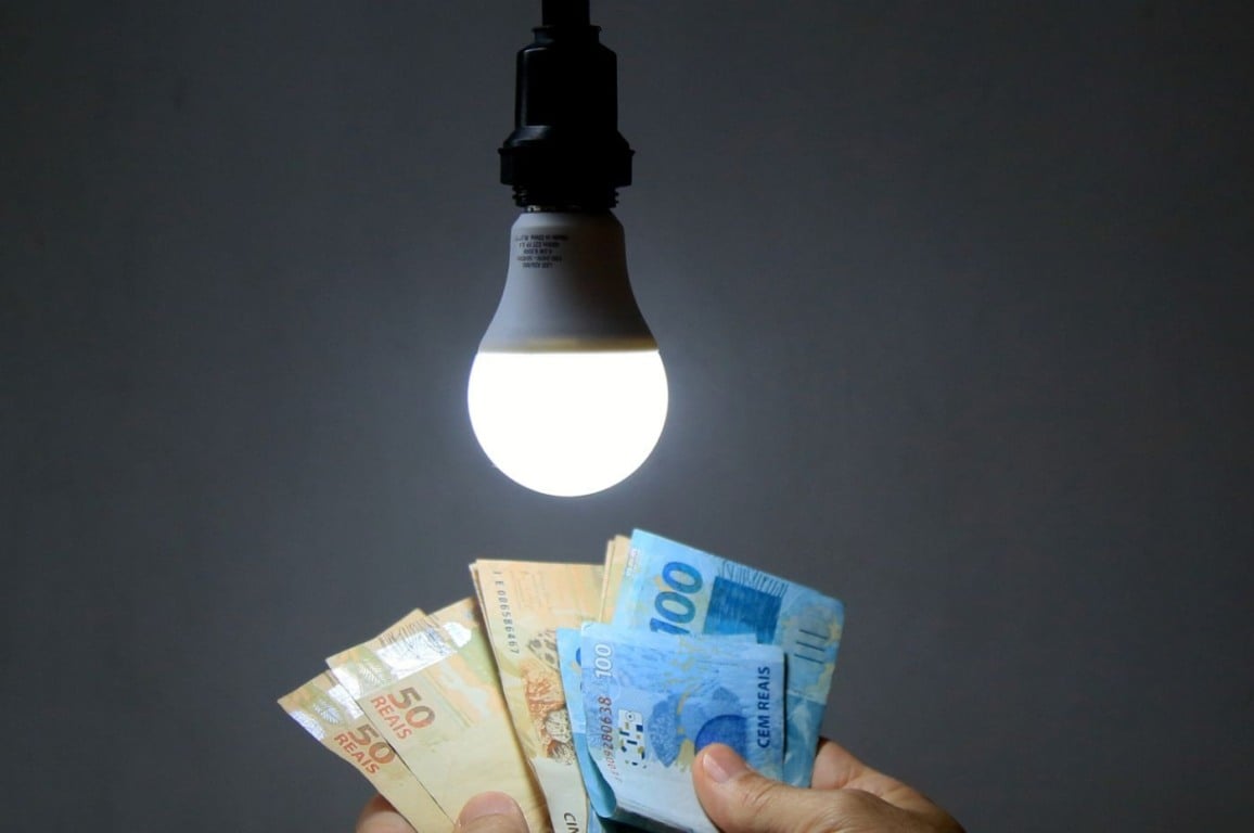 Conta de luz elevada, mãos segurando cédulas de real em frente a uma lâmpada acesa, simbolizando o aumento de tarifas.