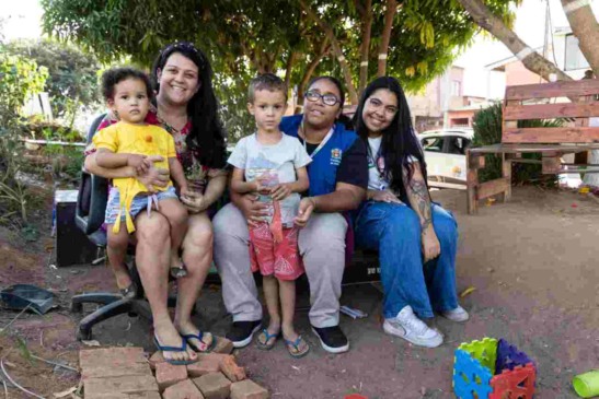 Grupo de pessoas sorrindo em um parque, com duas crianças ao centro e duas mulheres, uma delas usando uniforme do Programa Criança Feliz.