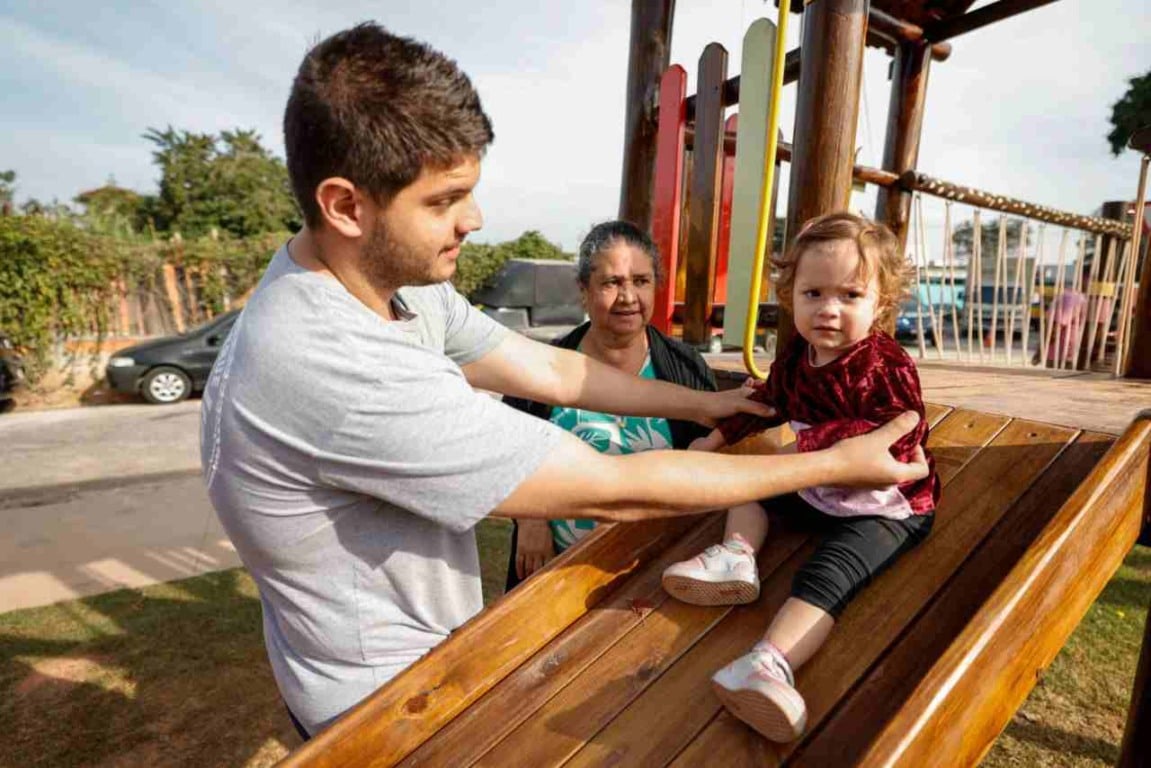 Pai ajudando filha pequena a descer escorregador com avó observando na Área da Infância em Jundiaí.