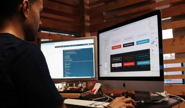 Designer trabalhando em dois monitores, desenvolvendo interface de usuário com códigos e botões em um ambiente moderno de escritório.