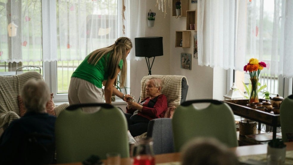 Cuidadora entrega bebida para idosa sentada em poltrona dentro de instituição de acolhimento. Cuidadora entrega bebida para idosa sentada em poltrona dentro de instituição de acolhimento.
