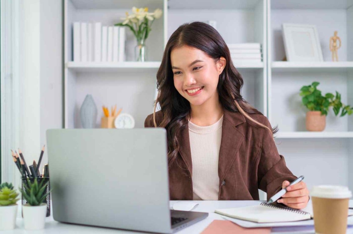 Mulher sorridente estudando em casa com laptop, caderno e café — curso gratuito em Jundiaí