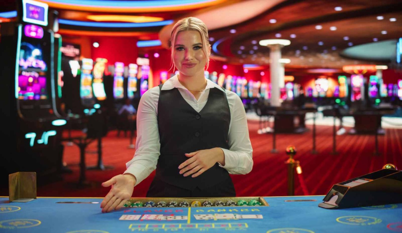 Dealer de cassino ao vivo sorrindo em uma mesa de jogo com cartas e fichas, cercada por máquinas caça-níqueis em um ambiente vibrante. Dealer de cassino ao vivo sorrindo em uma mesa de jogo com cartas e fichas, cercada por máquinas caça-níqueis em um ambiente vibrante.