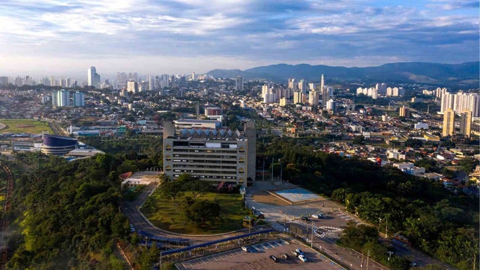 Vista aérea da cidade de Jundiaí com o prédio da Prefeitura Municipal em destaque, rodeado por áreas verdes, o Teatro Polytheama à esquerda e a paisagem urbana repleta de edifícios residenciais e comerciais ao fundo, sob céu parcialmente nublado.