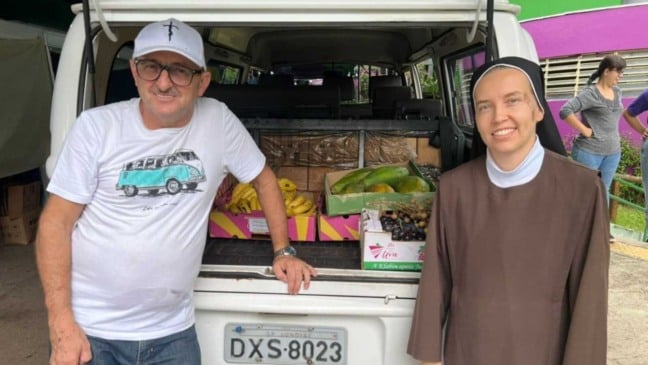 Homem e freira ao lado de veículo carregado de verdura e frutas.