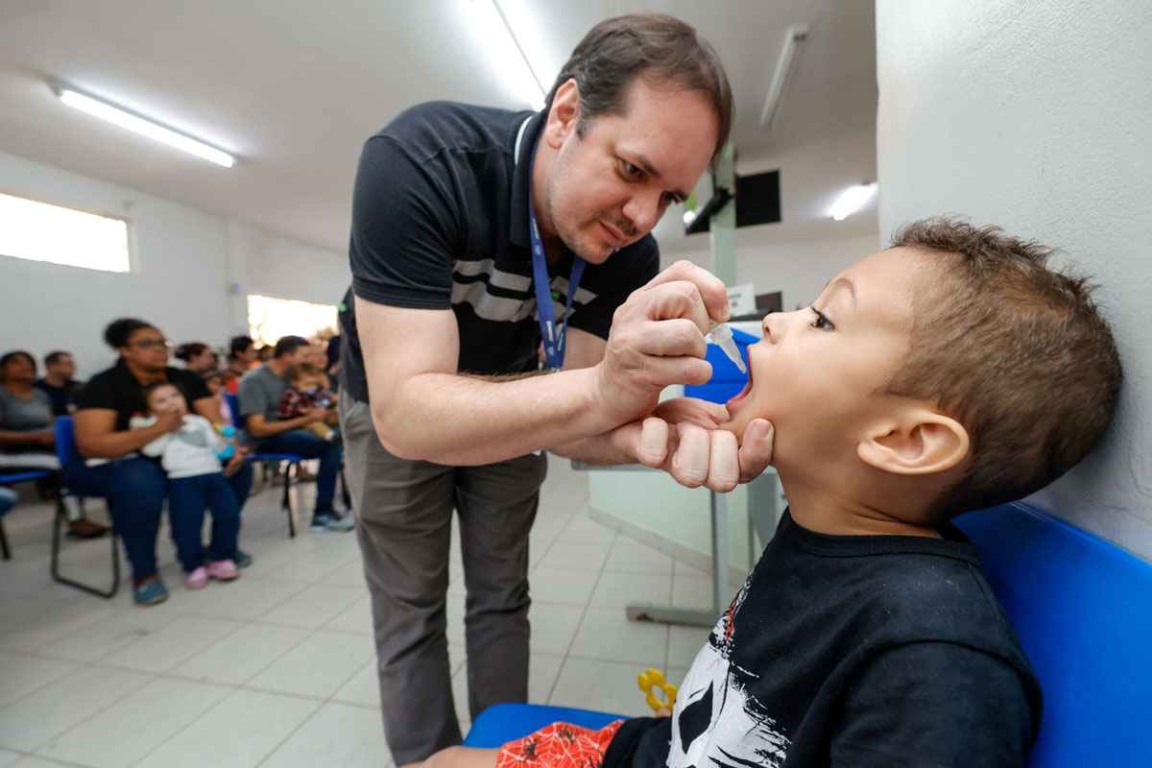 Profissional de saúde administra vacina oral contra poliomielite em uma criança, enquanto outras famílias aguardam na clínica.