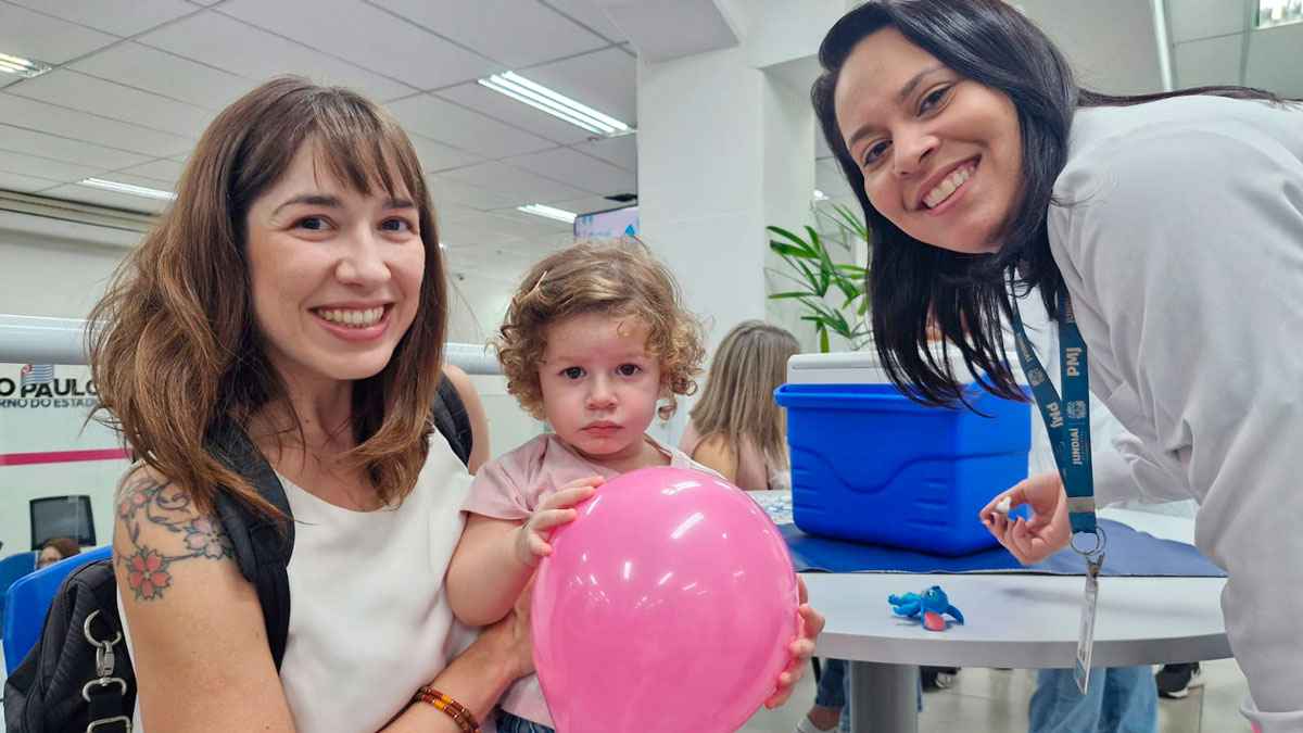 Mãe sorridente segura criança com balão rosa ao lado de profissional de saúde em um centro de vacinação, após receber vacina contra poliomielite.