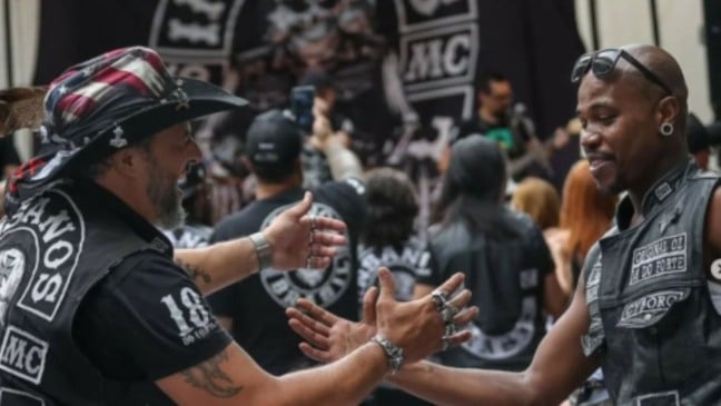 Dois homens em evento de motociclistas cumprimentando-se. Um usa chapéu com estampa da bandeira americana.