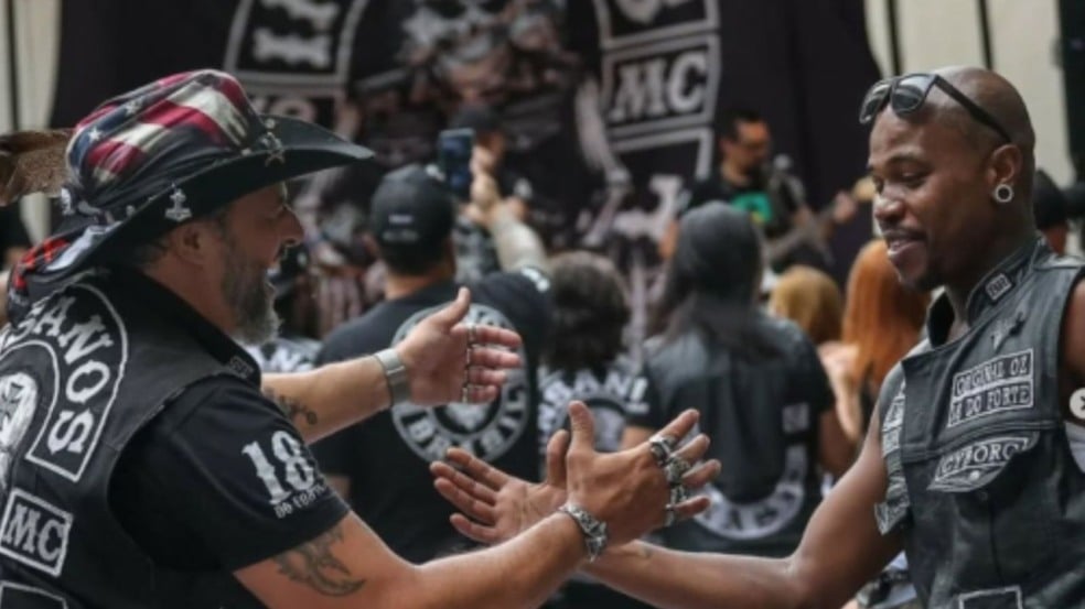 Dois homens em evento de motociclistas cumprimentando-se. Um usa chapéu com estampa da bandeira americana.