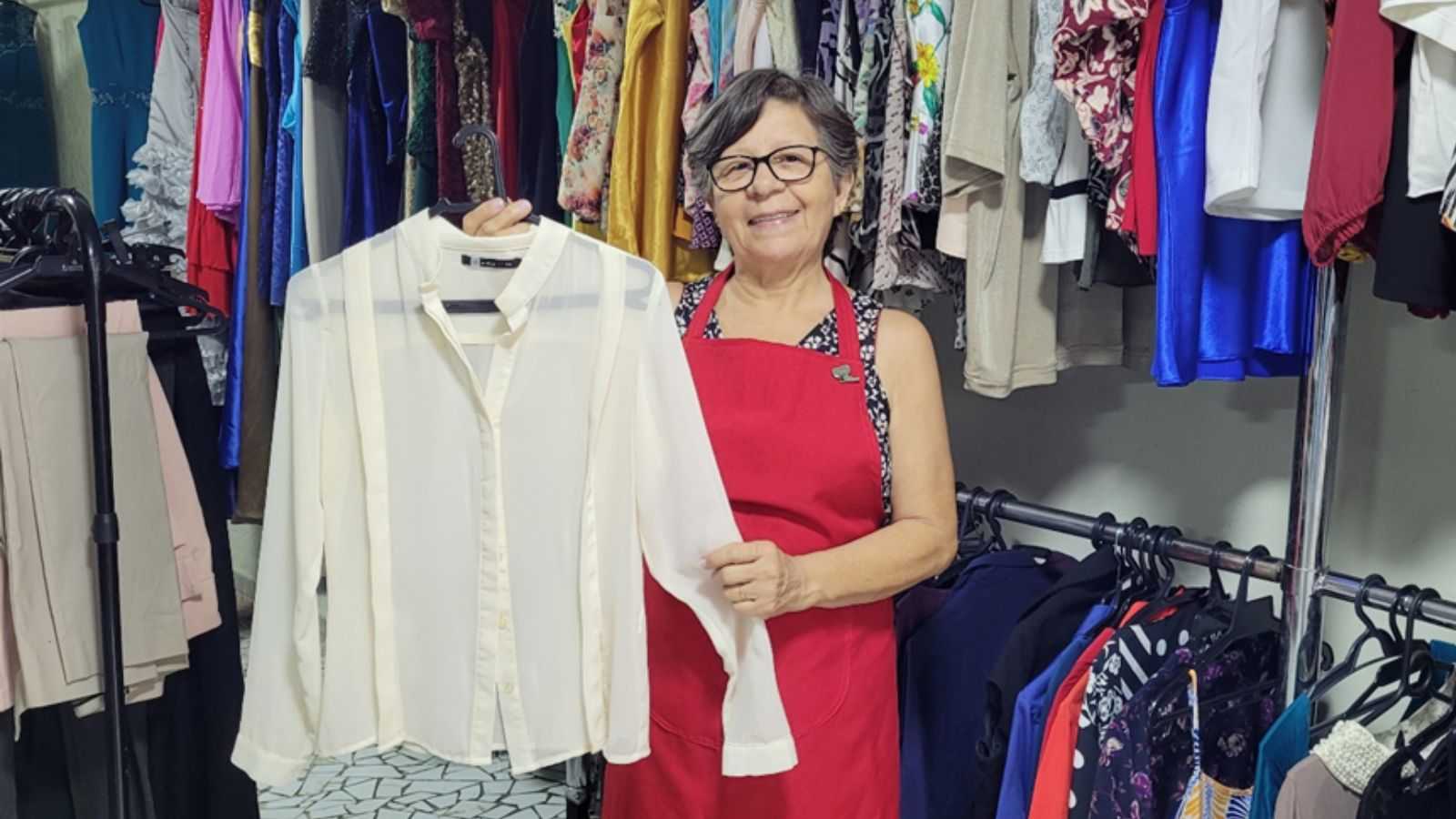 Voluntária do Hospital São Vicente em Jundiaí segura camisa branca em meio a roupas do Bazar do Voluntariado.