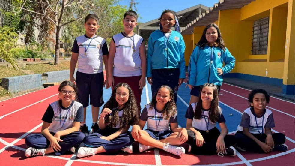 Crianças da EMEB Deodato Janski, em uniforme escolar, posam sorrindo em pista de atletismo com medalhas no pescoço após conquista na Olimpíada Internacional Matemática Sem Fronteiras 2025.