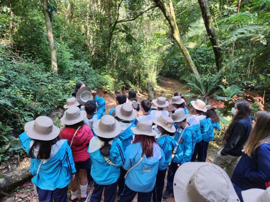 Um grupo de estudantes em uniformes azuis e chapéus bege está em uma trilha na Serra do Japi, participando de uma aula ao ar livre sobre mudanças climáticas.