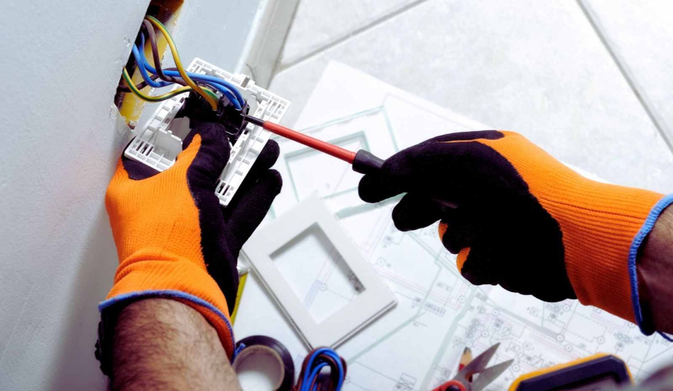 Curso de Eletricista Residencial do Fundo Social de Jundiaí: mãos com luvas laranja ajustando fios elétricos em uma tomada.