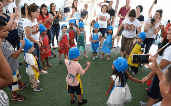 Crianças estão vestidas com chapéus e aventais, famílias estão ao redor
