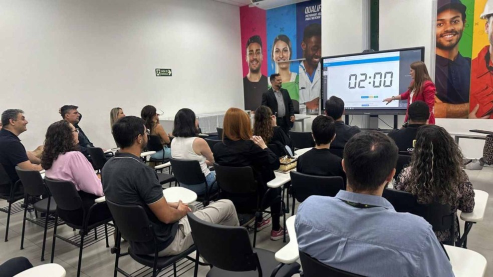 Sala de treinamento no Espaço Jundiaí Empreendedora com participantes sentados assistindo a uma apresentação; uma mulher de blazer rosa aponta para um telão digital com cronômetro de 2 minutos.