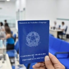 Pessoa segura uma Carteira de Trabalho e Previd&ecirc;ncia Social em destaque, com ao fundo sala de atendimento do Minist&eacute;rio do Trabalho.