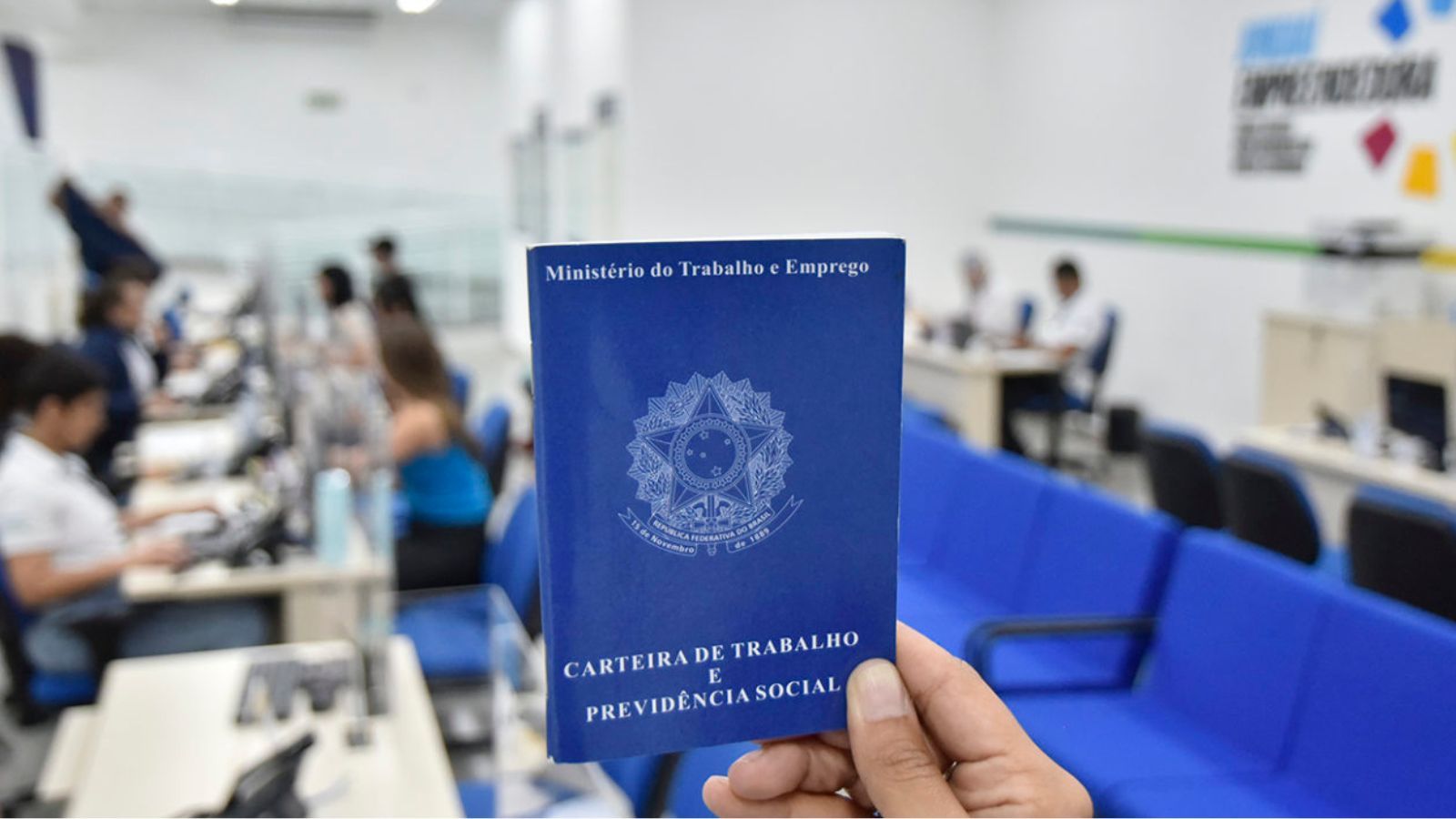 Pessoa segura uma Carteira de Trabalho e Previdência Social em destaque, com ao fundo sala de atendimento do Ministério do Trabalho.