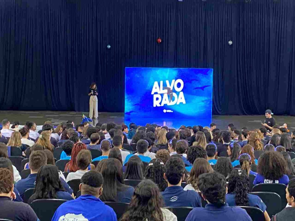 Palestrante se apresenta em um auditório cheio de pessoas, com uma tela grande ao fundo exibindo "Alvorada" em azul.