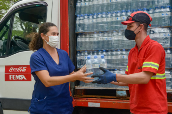 um homem com roupas da coca cola entregando um fardo de garrafas de água para uma mulher, com um caminhão atrás