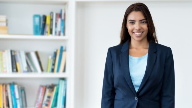 Mulher jovem sorridente usando blazer azul, em frente a uma estante de livros.