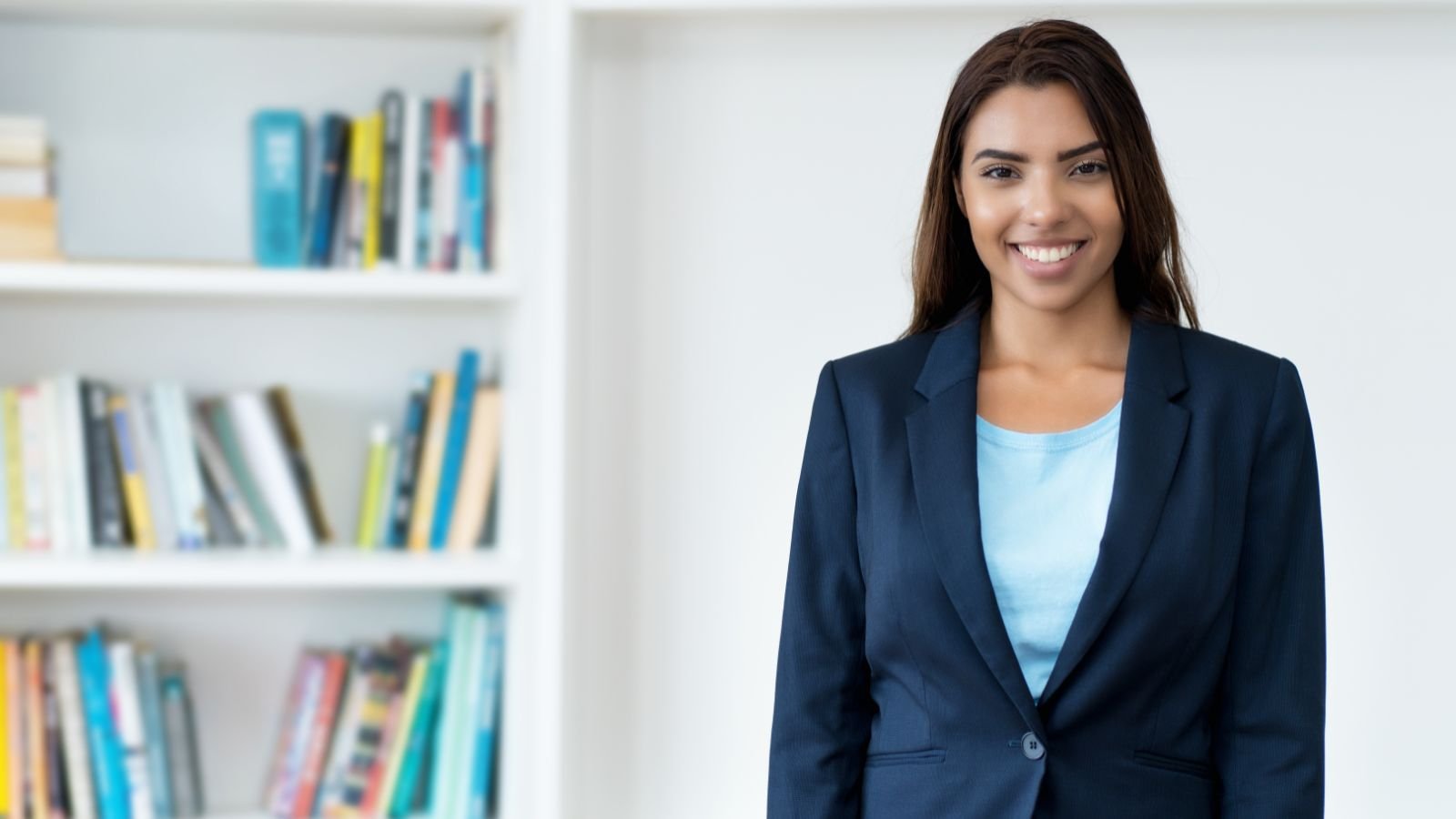 Mulher jovem sorridente usando blazer azul, em frente a uma estante de livros.