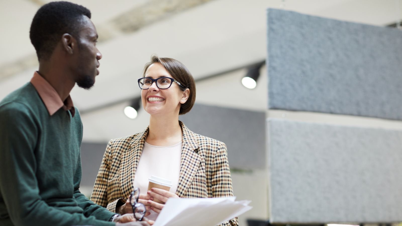 Dois colegas de trabalho conversando em ambiente corporativo, segurando documentos e café.