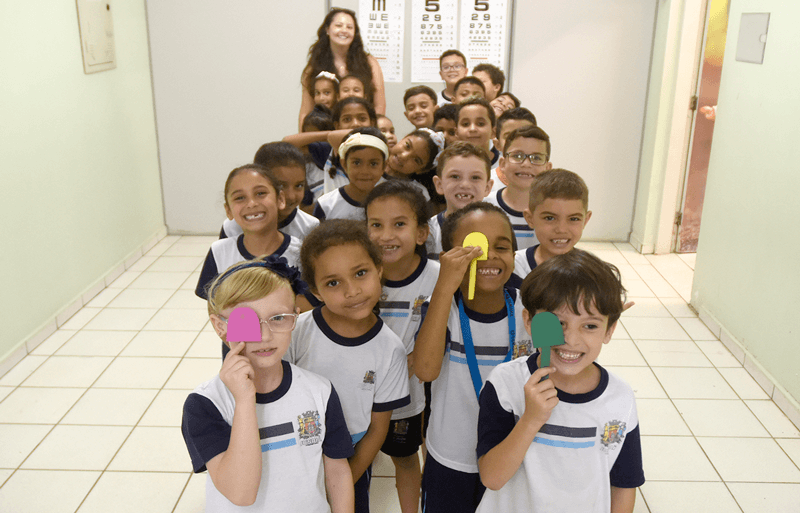 Crianças enfileiradas e professora no final da fila; todos utilizam ferramenta do teste para tampar um dos olhos