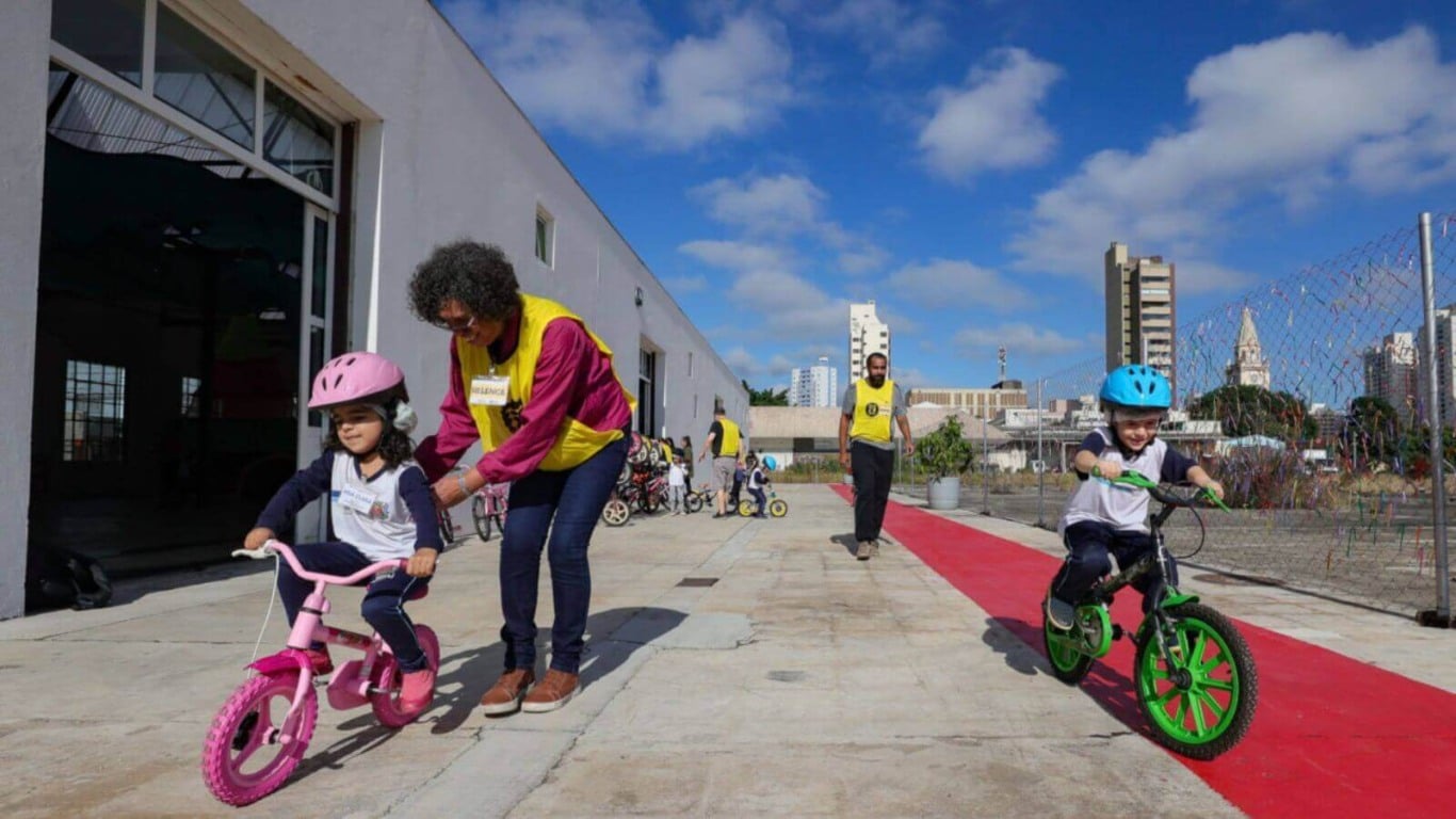 Crianças andando de bicicleta com capacetes, acompanhadas por monitores em um espaço ao ar livre, em frente a um prédio em Jundiaí.