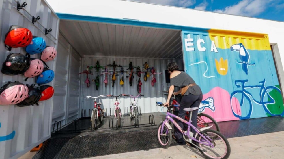 Imagem de uma mulher pegando uma bicicleta em uma bicicloteca de Jundiaí, que possui capacetes coloridos pendurados e bicicletas organizadas dentro de um contêiner decorado. Imagem de uma mulher pegando uma bicicleta em uma bicicloteca de Jundiaí, que possui capacetes coloridos pendurados e bicicletas organizadas dentro de um contêiner decorado.