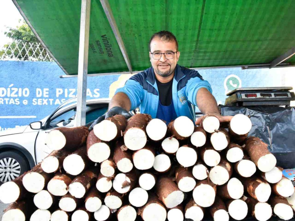 Homem sorridente, feirante regularizado, vendendo mandioca em uma barraca de feira em Jundiaí, com balança e cartaz ao fundo.