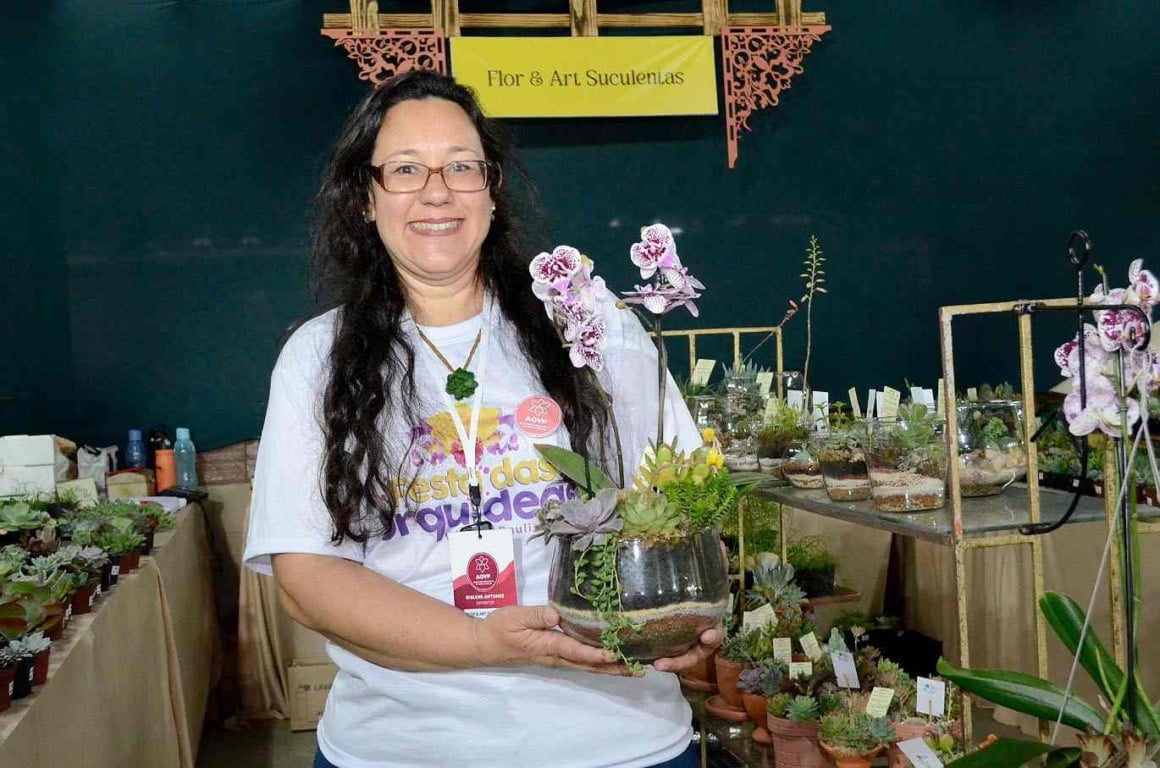 Expositora sorri enquanto segura orquídea na Festa das Orquídeas 2024, destacando o stand Flor & Art Suculentas em Várzea Paulista.