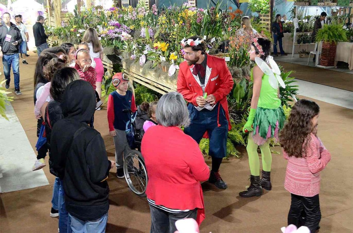Visitantes, incluindo crianças, interagem com personagens fantasiados na Festa das Orquídeas 2024, rodeados por flores em exposição.