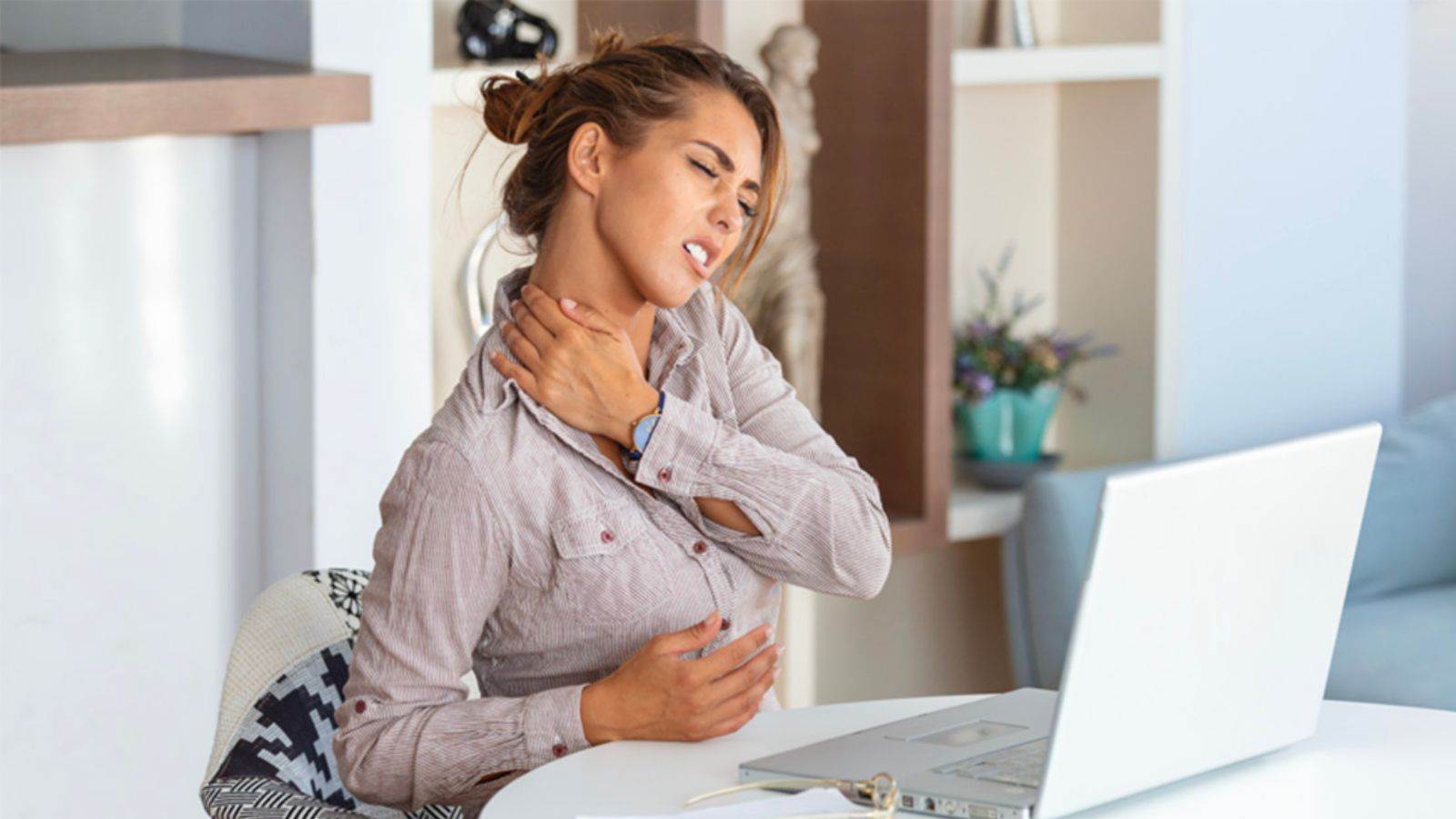 Mulher em frente ao notebook com expressão de dor, segurando o pescoço e o peito, ilustrando sintomas de fibromialgia.