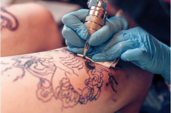 Pessoa recebendo uma tatuagem na perna feita por tatuador com luvas azuis, durante aplicação de desenho artístico detalhado.